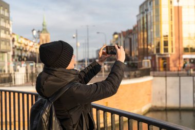 Young woman in black jacket and warm hat traveling with backpack, taking selfie on smartphone camera. Travel blogger photographing and record video message online. Tourism with gadgets concept.