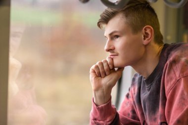Side view of young man in pink sweatshirt traveling by train and looks out of the window. Portrait of thoughtful blond man enjoying a train trip. Tourism and travel concept.