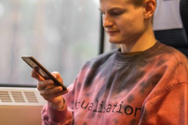 Young busy man in pink sweatshirt using smartphone when he traveling by train. Blond man sits at the train window, chatting in social media, uses mobile service and app. People and gadgets concept.