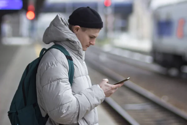 Side view of young man in casual gray jacket and hat using smartphone while waiting train at railway station. Travel blogger with backpack texting, cheks the route of trip on google mapes.
