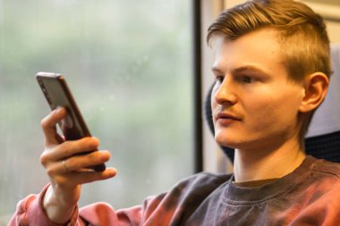 Young busy man in pink sweatshirt using smartphone when he traveling by train. Blond man sits at the train window, chatting in social media, uses mobile service and app. People and gadgets concept.
