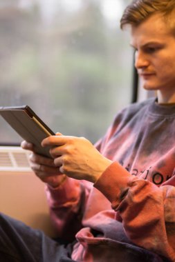 Smiling young man reading e-book when he traveling by train. Man sits at the train window and using digital tablet, watching movie, using mobile app, browsing internet, social media, shopping online.