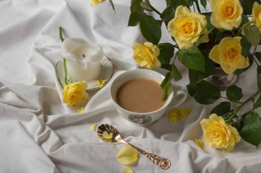Porselen kahve fincanı, klasik altın kaşık, bahçe sarısı güller, beyaz çarşafta mum. Ev konseptiyle iç içesin. Yatakta tatil kahvaltısı.