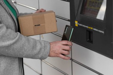 Pniewy, Polonya - 22 Mart 2023. Akıllı telefonu olan yaşlı bir kadın posta servisindeki alışveriş dolabını açmak ve paket göndermek için QR kodunu giriyor. E-ticaret nakliyat ve teslimat. İnsanlar ve aletler.