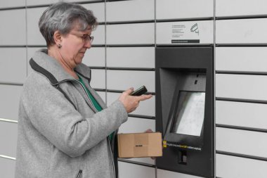 Pniewy, Polonya - 22 Mart 2023. Akıllı telefonu olan yaşlı bir kadın posta servisindeki alışveriş dolabını açmak için şifre giriyor ya da bir kutu gönderiyor. E-ticaret nakliye ve teslimat konsepti.