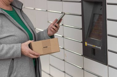 Pniewy, Polonya - 22 Mart 2023. Akıllı telefonu olan yaşlı bir kadın posta servisindeki alışveriş dolabını açmak için şifre giriyor ya da bir kutu gönderiyor. E-ticaret nakliye ve teslimat konsepti.