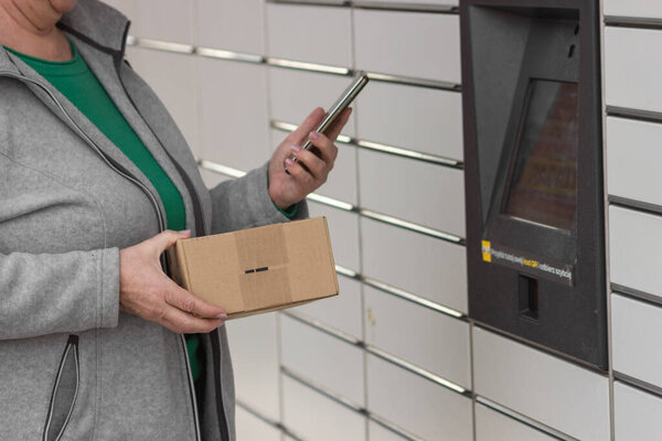 Pniewy, Poland - 22 March 2023. Senior woman with smartphone entering code to open shopping locker in inPost delivery service for receives or sends a box. E-commerce shipping and delivery concept.