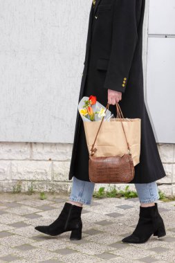 Siyah ceketli, çizmeli, deri çantalı ve çiçekli alışveriş çantalı zarif bir iş kadını. Alışveriş yaptıktan sonra caddede yürüyen şık bir kadın. Moda ayrıntılarını kapat.