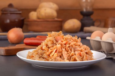Stewed cabbage - bigos in a white porcelain plate against the background of smoked sausages and chicken eggs. Close-up