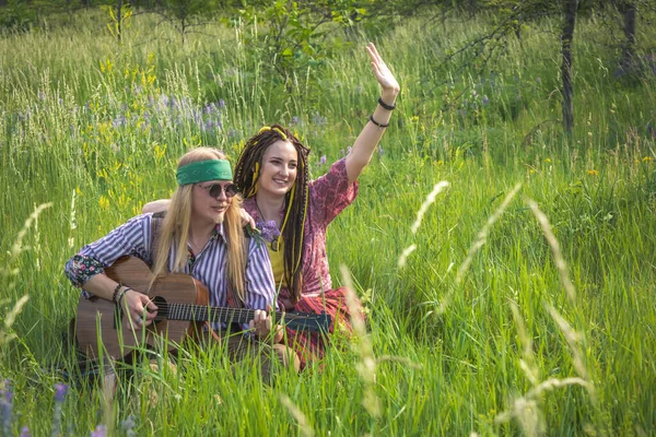 Hippie-style guy and a girl with a guitar are sitting in the grass on a forest glade on a sunny day and calling friends to join them