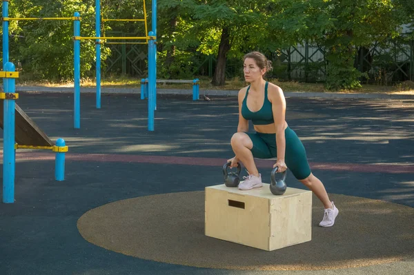Ellerinde kettlebell olan genç bir kadın, sokak spor sahasındaki tahta bir kutuda ayaklarıyla hamleler yapıyor. Sağlıklı yaşam tarzı
