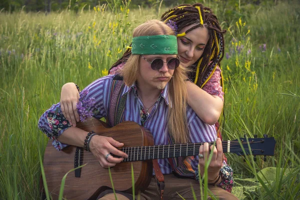 Guy and a girl in hippie style with a guitar are sitting in the grass on a forest glade on a sunny day. Close up