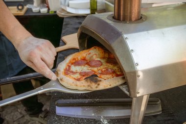 Fırıncı salamlı, peynirli ve domatesli pizzayı fırından çıkarmak için metal spatula kullanır. Yakın plan.
