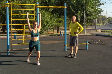 Bir sokak sporu sahasında lastik genişleticiyle egzersiz yapan bir eğitmenin rehberliğinde genç beyaz bir kadın.