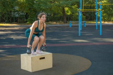 Elinde kettlebell olan genç bir kadın sokak sporları alanında tahta bir kutuda antrenman yapıyor. Sağlıklı yaşam tarzı