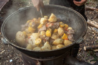Aşçı eti karıştırmak için slotlu kaşık, koçanda mısır, açık havada odun yakan bir kazanda patates ve sebzeler kullanır. Sokak yemeği.