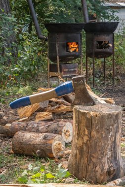 Balta, yakacak odunların arka planına karşı tahta bir bloğa saplanmış ve açık hava kazanları için yanan bir soba.