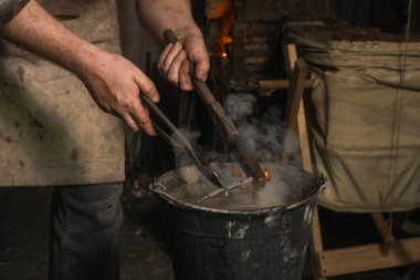 Demirci sıcak metal parçayı bir kova suda soğutur. Yakın plan.