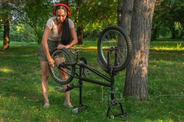 Büyük kırmızı kulaklıklı güzel Asyalı kız gün batımında parkta ya da ormanda ingiliz anahtarıyla bisiklet tamir ediyor. Sağlıklı yaşam tarzı.