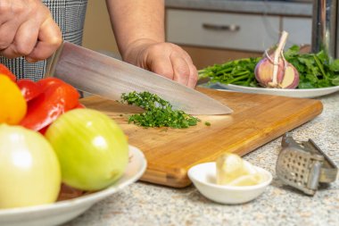 Mutfak tahtasındaki kadın aşçı çiğ sebzelerin arka planında salata için yeşillik kesiyor.