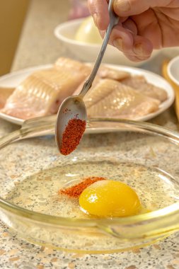 Cook adds ground paprika to a plate with a broken raw chicken egg to make liaison against the background of pieces of fresh fish and a bowl of flour. Close-up. Cooking at home clipart