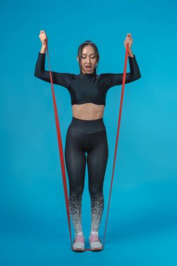 Slender Asyalı kız lastik genişletici ile spor giyim eğitimi alıyor. İzole edilmiş mavi arka plan. Sağlıklı yaşam tarzı kavramı.