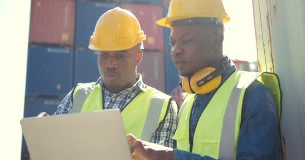 安全制服を着た2人のプロのアフリカ人エンジニアとラップトップで働くハード帽子 倉庫内で — ストック動画