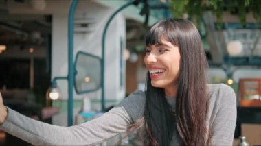 Genç bir kadın telefonda biriyle görüntülü görüşme yapıyor. Neşeli bir şekilde konuşuyor ve restoranda yüzünde bir gülümsemeyle.