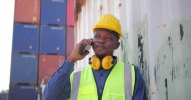 Lojistik kargo konteynırlarında cep telefonundan konuşan siyah Afrikalı işçi kaskı.