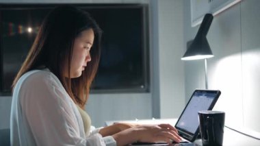 Asyalı bir kadın gece geç saatlere kadar çalışma odasında proje araştırması yaparak defter başında yazı yazıyor..