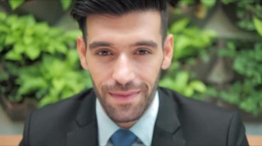 Zarif takım elbiseli gülümseyen profesyonel işadamı ofiste yeşil yapraklı kameraya bakıyor. Yüz görünümlü iş portresini kapat.