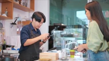 Asyalı kadın müşteri ve barista akıllı telefon kullanarak QR kod etiketini tarıyor. Kahve dükkanında veya restoranda kahve içen başka bir akıllı, para almadan dijital ödeme yapmayı kabul ediyor..