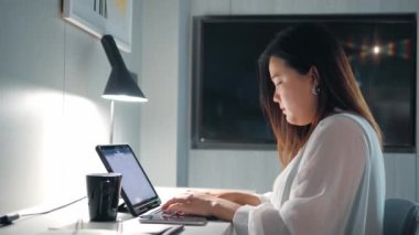 Asyalı bir kadın masa başında oturmuş klavyeyle yazı yazıyor ve gece geç saatlere kadar odada proje araştırması yapıyor..
