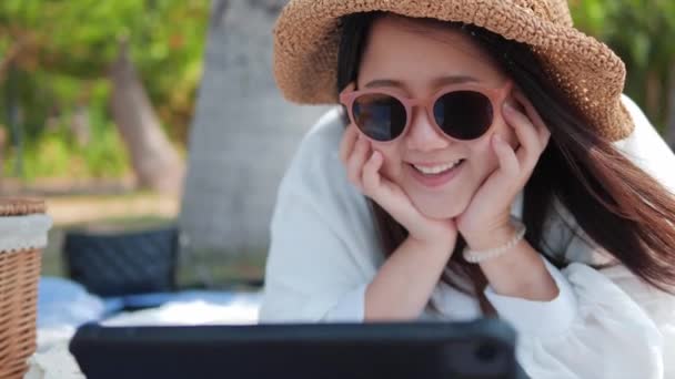 ビーチパークの芝生の上に帽子と眼鏡をかぶったアジアの女性は タブレットでビデオを見て 夏の週末にリラックス笑って — ストック動画