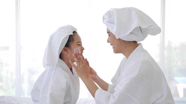 アジアのお母さんAdding治療クリーム上のザ頬に女の子にスパドレスと頭カバーでAホワイトタオルでベッド一緒に家 — ストック写真