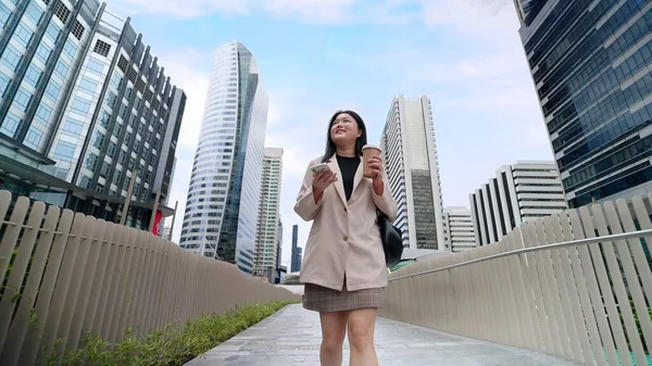 スマートフォンやコーヒーカップを持ってスーツを着て街の中心部で街の景色を見て回るアジアのビジネスマン女性 — ストック写真