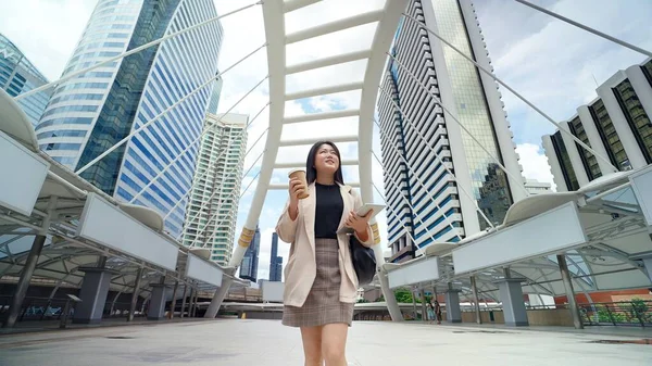 スーツを着たアジアの実業家がタブレットとコーヒーカップを持って街の中心部を見て通りを歩いている — ストック写真