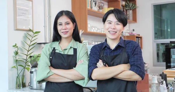 A couple of Asian women have a smiling face and wearing an apron standing in front of the counter In a coffee shop. The Lesbian and tomboy couple owns and operates a cafe together