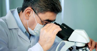 Yüz maskesi takan Asyalı doktor, aşı örneğini laboratuvar ve aşı araştırmalarında mikroskop ile analiz ediyor. Karantina veya virüs salgını kavramı