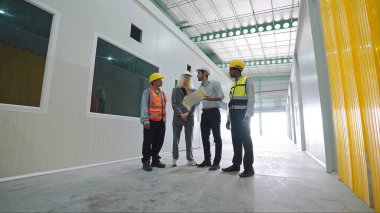 Bir grup fabrika çalışanı depoda dikilip iş hakkında konuşuyorlar. Depoda güvenlik kaskı denetimi yapıyorlar.