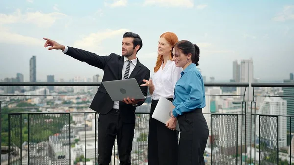 A team of business people standing, talking and using laptop to work at rooftop city view