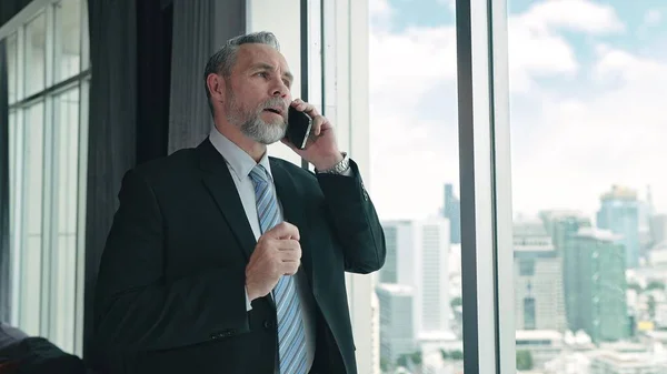オフィスの大きなガラス窓の近くに立っているスマートフォンの上で話をしているスーツを着ている上級ビジネスマン — ストック写真