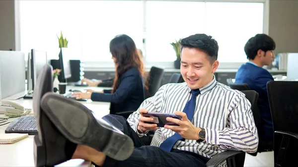 楽しいゲームをしている怠惰なアジアのビジネスマンは 電話でオフィスで働く時間を費やして 同僚は義務を果たします — ストック写真