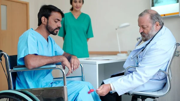 病院の治療室で車椅子に座っているインドの男性患者に膝の痛みを治療するアジアの高齢男性医師 — ストック写真