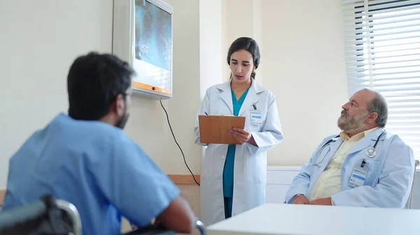 女性医療アシスタントは クリップボード上の治療情報を書きますシニア男性医師とインドの男性患者がそこに座って説明結果を聞きます — ストック写真