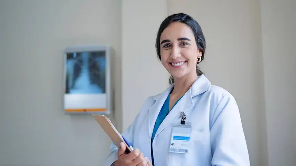 笑顔若いですヒスパニックラテン女性医師でホワイト医療コート保持クリップボード上の立っていると見ますカメラで病院 — ストック写真