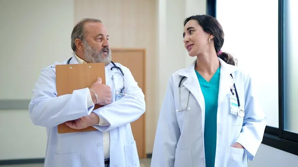 病院の廊下を歩いている間に診断を議論する2人の医者は白いコートを身に着けている 若いです女性医師と話すシニア男性医師監督 — ストック写真