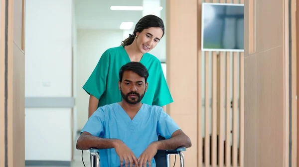 インド人男性患者の車椅子を押して病院の治療室に行く若い女性看護師 — ストック写真