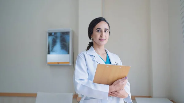 若いですヒスパニックラテン女性医師でホワイト医療コート保持クリップボード上の立っていると見ますカメラで病院 — ストック写真