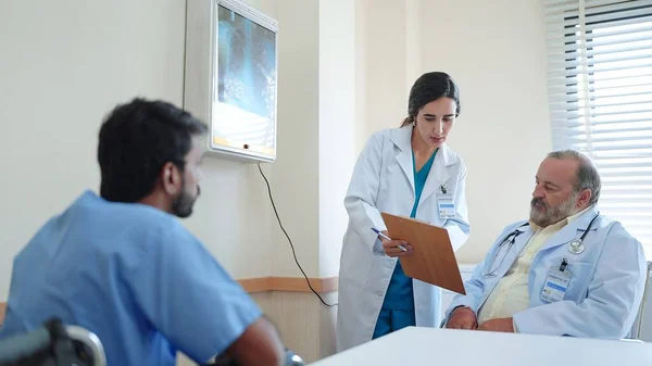女性医療アシスタントは クリップボード上の治療情報を書きますシニア男性医師とインドの男性患者がそこに座って説明結果を聞きます — ストック写真
